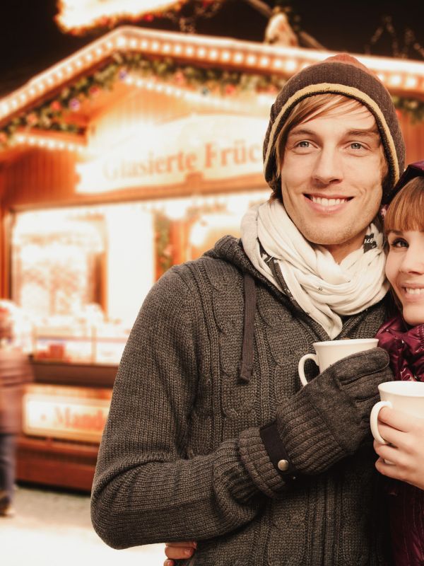 Ein Paar genießt warme Getränke vor einem festlich beleuchteten Weihnachtsmarktstand.